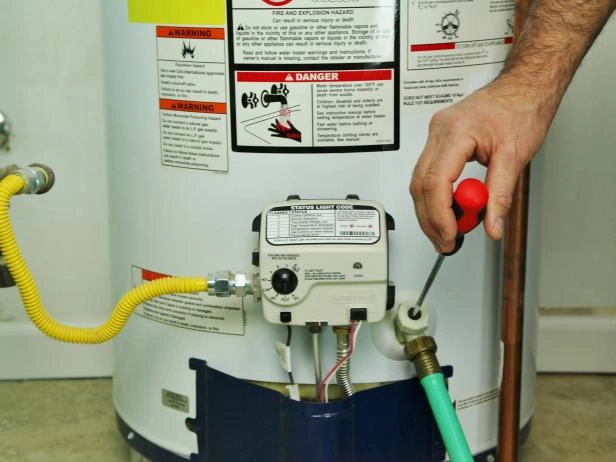 Person adjusting a gas water heater thermostat with a screwdriver. Warning labels visible.