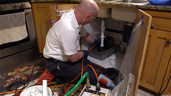 A plumber kneeling and working under a kitchen sink.