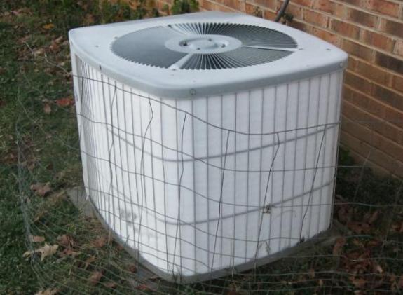 An outdoor air conditioning unit with a protective wire cage against a brick wall.
