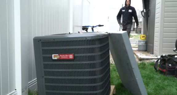 Outdoor AC unit in focus with a person blurred in the background near a house.