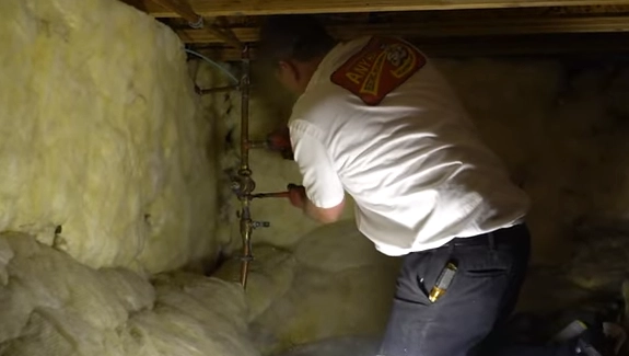 A person inspecting or working on pipes in a crawlspace with insulation.