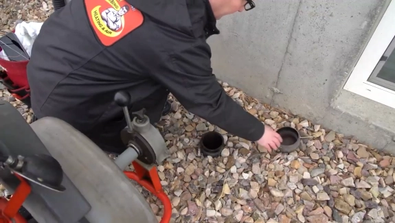 Person in a black jacket working on an outdoor pipe next to a wall.