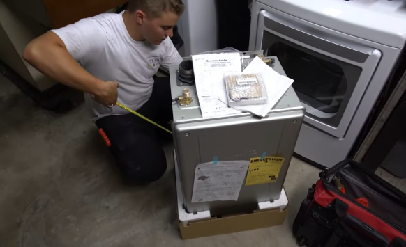 Person measuring a tankless water heater next to an laundry machine in a dimly lit room.