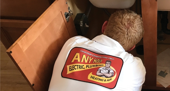 A worker fixing a sink, seen from behind, with a company logo on the back of his shirt.