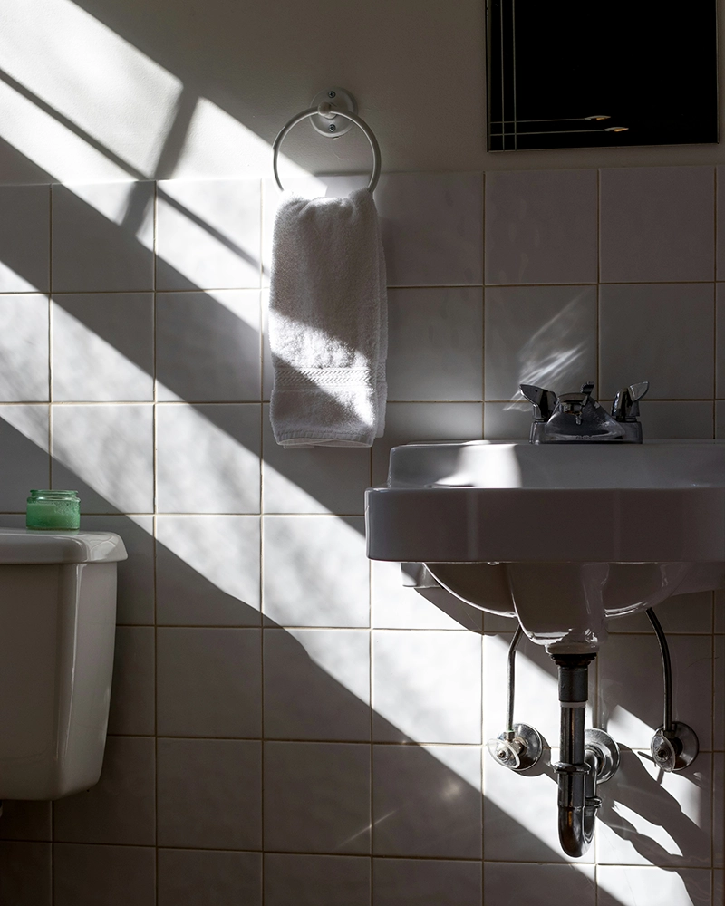 A sunlit bathroom with a white towel hanging, basin, faucet, and toilet against tiled walls.