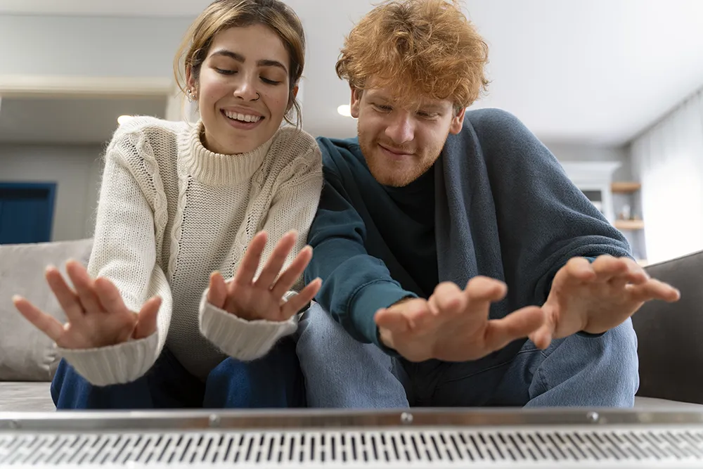 A smiling woman and a man, both dressed in cozy sweaters, sit on a couch and warm their hands over a heater in a bright, comfortable living room.