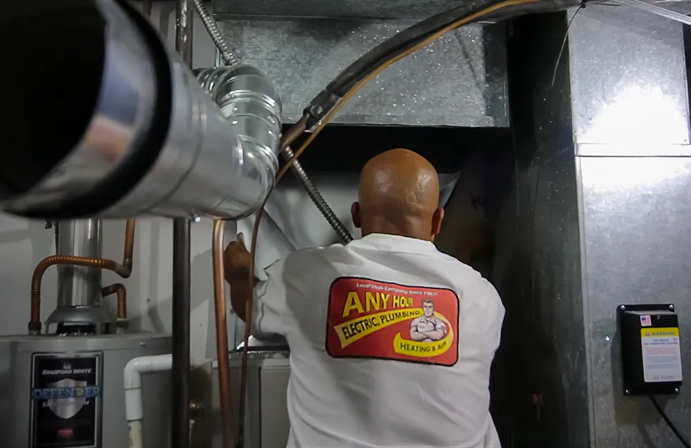 A person in a work uniform labeled Any Hour is inspecting a metal HVAC system in a utility room. The scene conveys professionalism and focus.